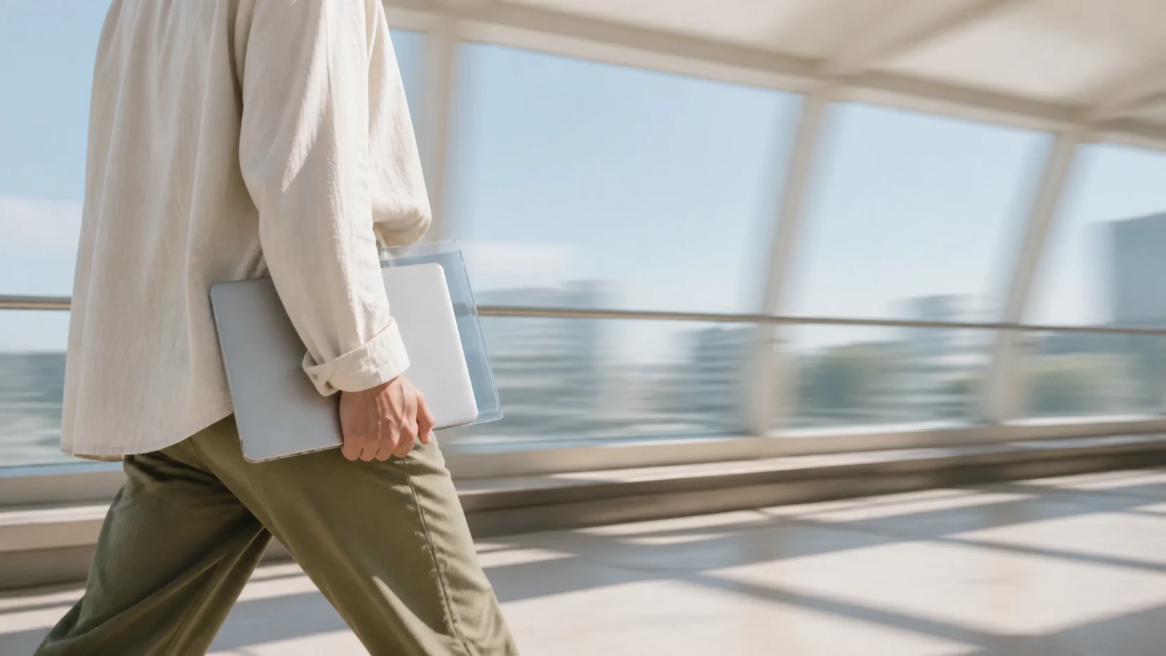 A person carrying a laptop and translucent prompt library sleeve through a bright open walkway